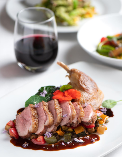 A plate of sliced duck breast on a bed of colorful vegetables, garnished with flowers, alongside a glass of red wine.